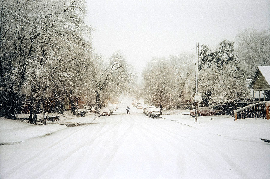 A picture of a winter storm-the roads, trees, and buildings all covered in a thick coat of snow. A loan individual stands in the middle of the road.